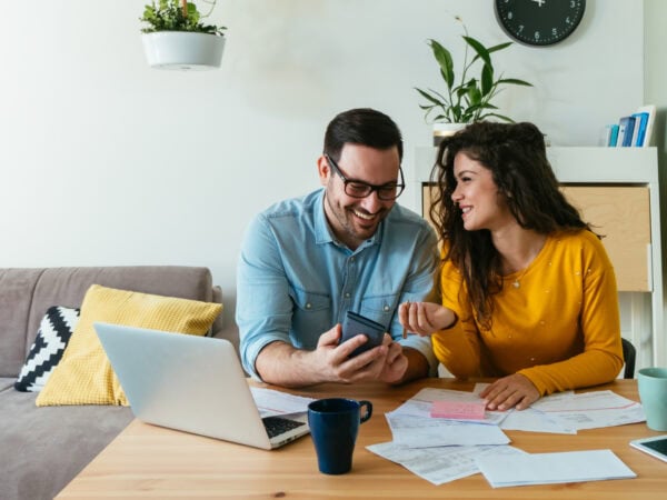 Smiling boyfriend showing calculator to his cheerful girlfriend while doing home finances together online on a laptop computer in the living room.